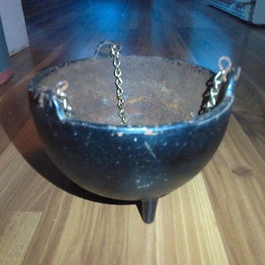 May include: A black cast iron cauldron with three small legs and a gold chain handle. The cauldron has a rounded shape and a rustic, aged appearance, suggesting it may be used for cooking or decorative purposes.