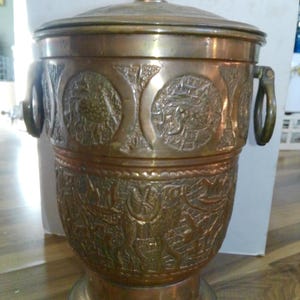 May include: A cylindrical brass ice bucket with a lid and two side ring handles. The exterior features embossed circular panels depicting animal figures and intricate patterns. The metal has a warm, aged patina with visible texture.