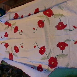 May include: White quilt with red poppy flower embroidery and green leaves. The quilt has a scalloped red border. The floral design is spread across the quilt's surface. The quilt is folded and appears to be on a bed.