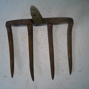 May include: An antique, rusty metal garden tool with four tines and a central spike. The tool has a weathered appearance, suggesting age and use. The handle is attached to the top of the tool.