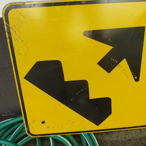 May include: Yellow road sign with black arrows indicating a change in elevation. The sign has a weathered appearance, with scratches and wear. The sign is rectangular with rounded corners.