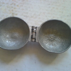 May include: A silver metal egg cracker with a hinged design. The cracker has two halves, each with a textured surface.