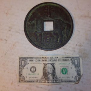 May include: A round, bronze-colored coin with a square hole in the center, featuring a horse design. A one-dollar bill is placed below the coin for size comparison.