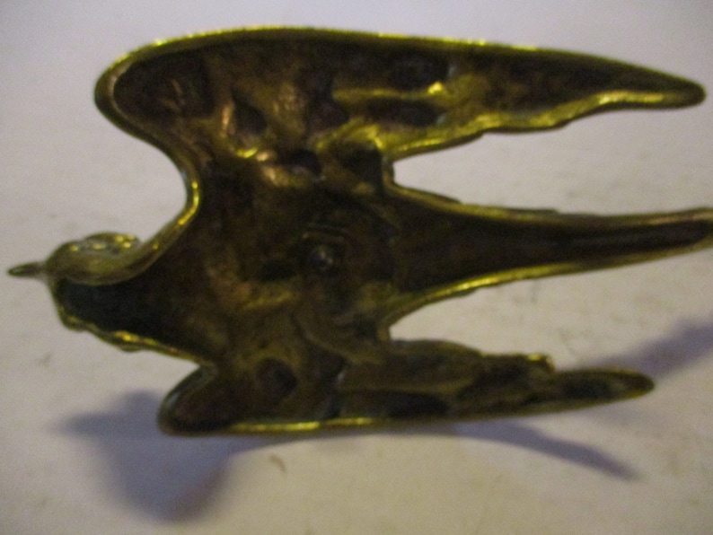 K&ouml;nnte beinhalten: Vintage Vogel-Figur aus Messing, vermutlich ein K&uuml;hlerfigur. Der Vogel wird im Flug mit ausgebreiteten Fl&uuml;geln dargestellt. Das Messing hat eine warme, gealterte Patina, die auf sein Alter hindeutet. Die Skulptur ist detailliert und zeigt die Federn und Form des Vogels.