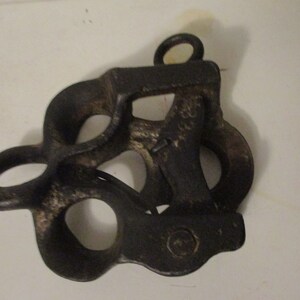 May include: A black cast iron pulley with multiple loops and a single hook. The pulley has a worn, aged appearance.