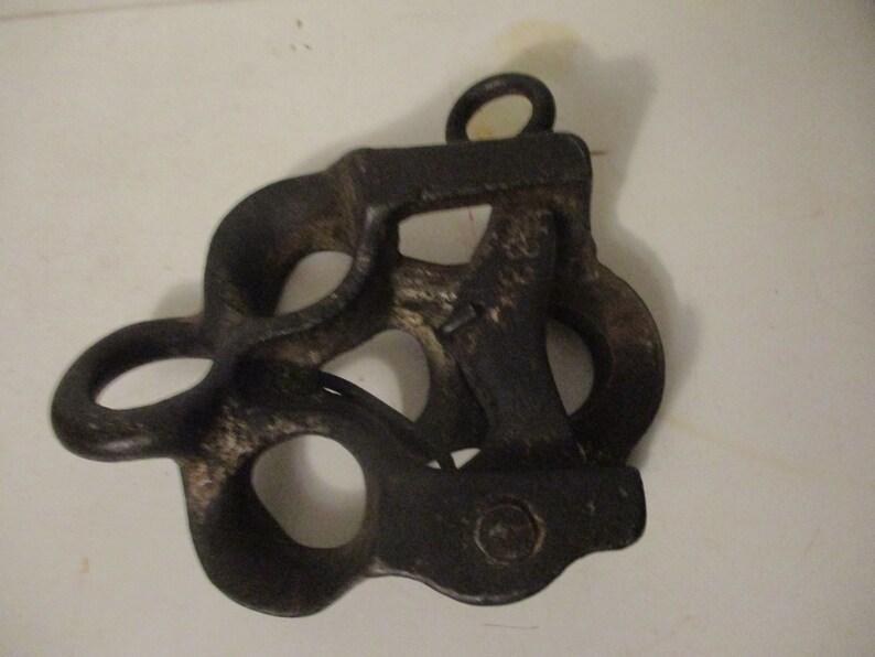 May include: A black cast iron pulley with multiple loops and a single hook. The pulley has a worn, aged appearance.