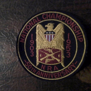 May include: A black and gold embroidered patch with the text "National Championship 1903-1953 50th Anniversary NRA". The patch features a golden eagle with a red, white, and blue shield and two crossed rifles.