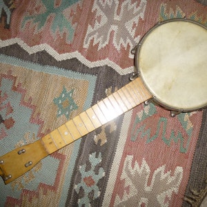 May include: A vintage banjo with a round, cream-colored drumhead and a light-colored wooden neck. The banjo has metal hardware and is set against a patterned rug with shades of brown, blue, and pink.