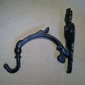 May include: Two black cast iron plant hangers. One is a wall-mounted bracket with a decorative bird design. The other is a curved arm with a hook. Both are designed to hold hanging baskets or other items.