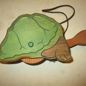 May include: A wooden turtle toy with a green shell and brown head and feet. The shell has a leaf-like design. A brown string is attached to the top. The toy is painted with a light green and brown color scheme.