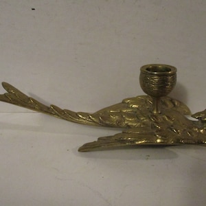 K&ouml;nnte beinhalten: Ein messingfarbener Vogel-Kerzenhalter. Der Vogel wird im Flug dargestellt, mit ausgebreiteten Fl&uuml;geln und einem detaillierten Federdesign. Ein kleiner, becherf&ouml;rmiger Halter sitzt auf dem R&uuml;cken des Vogels. Der Kerzenhalter ist aus Messing und hat eine gl&auml;nzende Oberfl&auml;che.