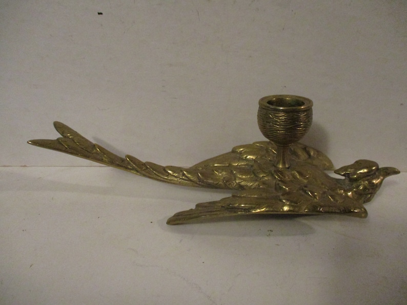 K&ouml;nnte beinhalten: Ein messingfarbener Vogel-Kerzenhalter. Der Vogel wird im Flug dargestellt, mit ausgebreiteten Fl&uuml;geln und einem detaillierten Federdesign. Ein kleiner, becherf&ouml;rmiger Halter sitzt auf dem R&uuml;cken des Vogels. Der Kerzenhalter ist aus Messing und hat eine gl&auml;nzende Oberfl&auml;che.