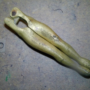 May include: A vintage, gold-colored metal tool with a unique, curved design. The tool has two elongated, leg-like arms connected at the top, with a small hole. The surface shows signs of wear.