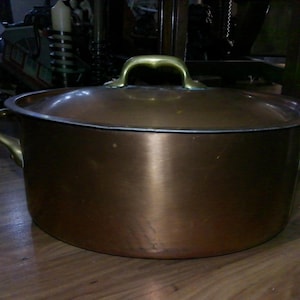May include: A copper-colored cooking pot with a matching lid. The pot has two side handles and a brass handle on the lid. The pot is sitting on a wooden surface.