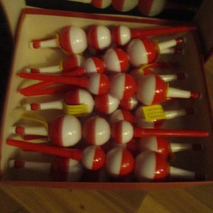 May include: A box of vintage fishing bobbers. Each bobber is white and red, with a long red stem. The bobbers are arranged neatly inside a cardboard box, suggesting they are new or unused.