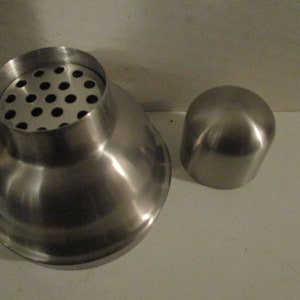 K&ouml;nnte beinhalten: Ein Cocktailshaker aus Edelstahl mit einem Sieb und einem separaten Deckel. Der Shaker hat eine polierte, reflektierende Oberfl&auml;che. Das Sieb hat mehrere kleine L&ouml;cher. Der Deckel ist abgerundet.