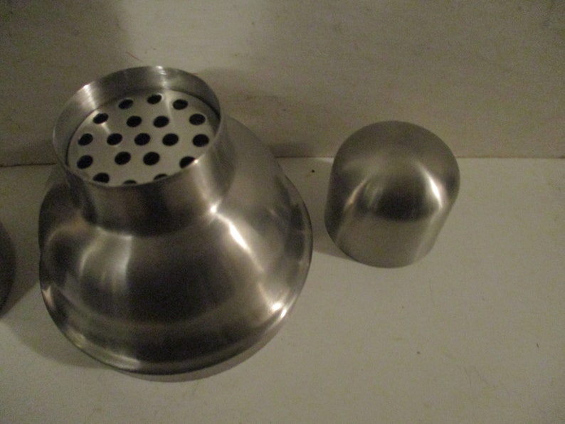 K&ouml;nnte beinhalten: Ein Cocktailshaker aus Edelstahl mit einem Sieb und einem separaten Deckel. Der Shaker hat eine polierte, reflektierende Oberfl&auml;che. Das Sieb hat mehrere kleine L&ouml;cher. Der Deckel ist abgerundet.