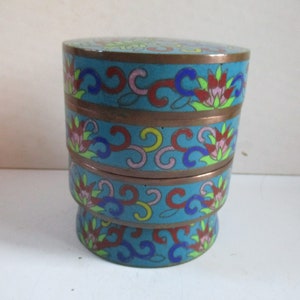 May include: A stacked, round, decorative box with a turquoise background and floral patterns in red, pink, yellow, and blue. The box has bronze-colored bands separating the sections. The overall design is ornate and detailed.