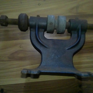 May include: A vintage metal lathe tool with a dark, aged appearance. The tool features a bolt, a series of cylindrical components, and a U-shaped base. The metal has a weathered look, suggesting age and use. The tool is designed for woodworking or metalworking.