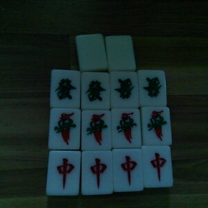 Pu&ograve; includere: Una collezione di quattordici tessere rettangolari bianche da mahjong disposte in una griglia. Le tessere presentano simboli incisi in rosso e verde, inclusi caratteri cinesi e motivi floreali, con due tessere vuote posizionate in alto.