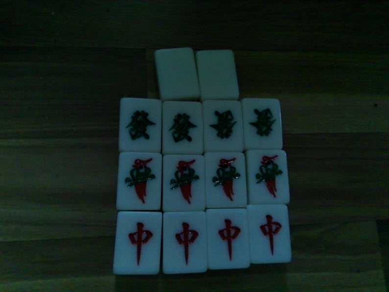 Pu&ograve; includere: Una collezione di quattordici tessere rettangolari bianche da mahjong disposte in una griglia. Le tessere presentano simboli incisi in rosso e verde, inclusi caratteri cinesi e motivi floreali, con due tessere vuote posizionate in alto.