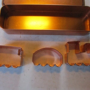 May include: Copper-colored cookie cutters in the shape of a train and a train car, along with a rectangular container. The train shapes include a locomotive and a caboose. The set is made of shiny copper-colored metal.