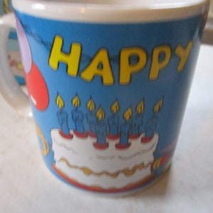 Puede incluir: Una taza de cer&aacute;mica azul con un borde y un asa blancos. La taza tiene un tema de cumplea&ntilde;os con las palabras "HAPPY" en letras amarillas, un pastel con velas azules y globos rojos y morados.