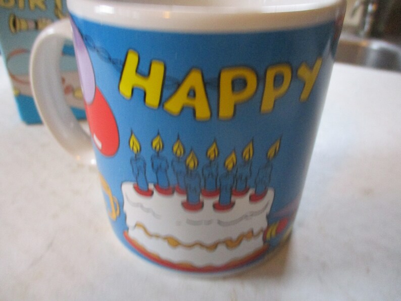 Puede incluir: Una taza de cer&aacute;mica azul con un borde y un asa blancos. La taza tiene un tema de cumplea&ntilde;os con las palabras "HAPPY" en letras amarillas, un pastel con velas azules y globos rojos y morados.