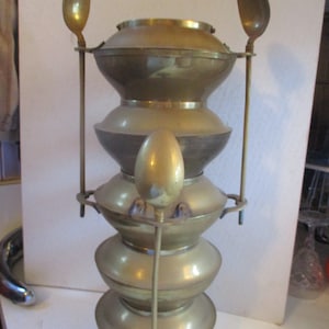 May include: A vintage brass serving piece with a tiered design. The piece has a series of rounded, stacked sections, with two spoons attached on either side. A central rod supports an egg-shaped finial.