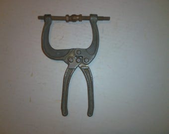 May include: A silver metal C-clamp with a threaded rod and a handle. The clamp is used for holding objects together.