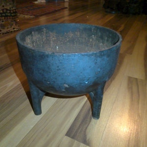 May include: A black cast iron cauldron with three legs. The cauldron is sitting on a wooden floor.