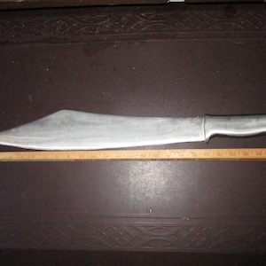 May include: A large, silver-colored machete with a thick blade and a sturdy handle. The blade is approximately 18 inches long. A wooden ruler is placed next to the machete for scale, showing the blade's length.