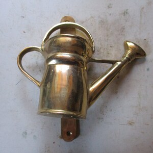 May include: A brass watering can wall decor with a wooden base. The watering can has a spout and a handle.