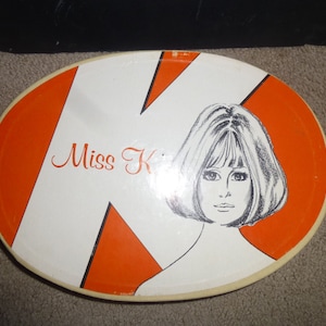 May include: Oval serving tray with a retro design. The tray features a white background with orange accents and the words "Miss K" in cursive. A black and white illustration of a woman's face is also on the tray.