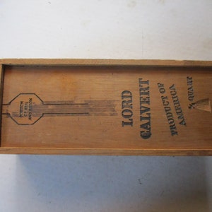 May include: Wooden box with the text "LORD CALVERT PRODUCT OF AMERICA" printed in black. The box also has a seal and a measurement of 4/5 Quart. The box is made of light brown wood.