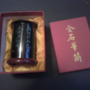 May include: A black cylindrical vase with gold Chinese characters on the side, inside a red box with a gold satin lining. The box has gold Chinese characters on the top that read "Golden Unbreakable Harmony".
