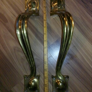 May include: Two antique brass door handles with a curved design. Each handle has a square base and a decorative top. A wooden ruler is placed between the handles, showing a measurement of approximately 14 inches.