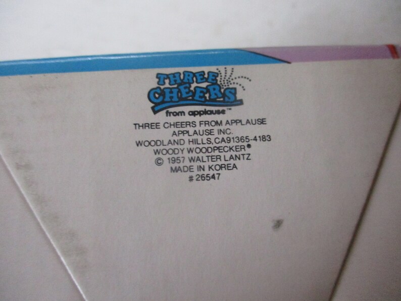 Puede incluir: Una caja blanca con bordes azules y morados. La caja tiene texto que dice "Three Cheers from Applause" y "Woody Woodpecker". La caja tambi&eacute;n incluye el texto "Three Cheers from Applause, Applause Inc., Woodland Hills, CA 91365-4183, &copy; 1957 Walter Lantz, Made in Korea, #26547".