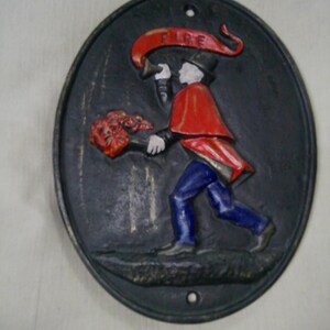 May include: Oval, cast iron wall plaque featuring a firefighter in a red cape and blue pants, holding a bouquet and a horn. The word "FIRE" is on a banner above the figure. The plaque is painted in black, red, blue, and white.