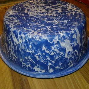 May include: A vintage enamelware bowl with a distinctive blue and white marbled pattern. The bowl has a wide, shallow design with a rounded base and a flared rim. The blue and white pattern gives it a classic, rustic look.