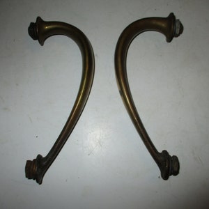 May include: Two antique brass-toned cabinet handles. Each handle has a curved, ornate design with a flared base and a decorative top. The handles are likely for a drawer or cabinet door.
