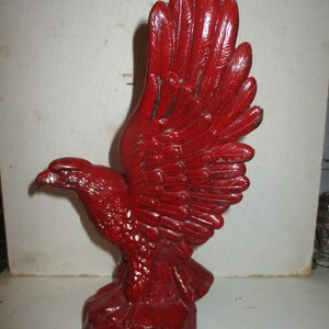 May include: A red painted eagle statue with outstretched wings. The sculpture has detailed feathering and is mounted on a rocky base. The eagle is a symbol of freedom and strength.