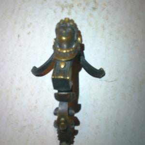 May include: A gold-toned metal decorative stand with a human figure head at the top. The stand has a three-pronged base and intricate details.