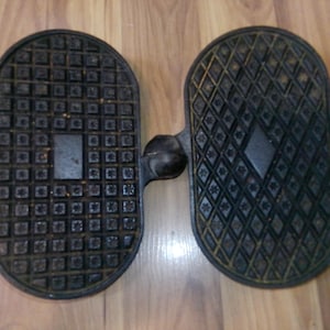 May include: Two black, textured foot pedals connected by a metal bar. One pedal has a square grid pattern, the other a diamond pattern. The pedals are likely part of an exercise machine or vintage equipment.