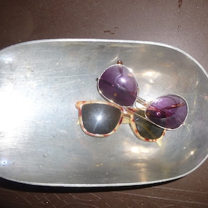 May include: Two pairs of sunglasses rest in a silver metal scoop. One pair has a tortoiseshell pattern with black lenses, and the other has purple lenses and a silver frame. The scoop has a handle.