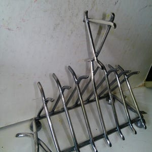 May include: A chrome-plated metal toast rack with a rectangular base and seven vertical slots. The rack has a handle at the top and curved feet. The design is simple and functional, suitable for holding slices of bread.