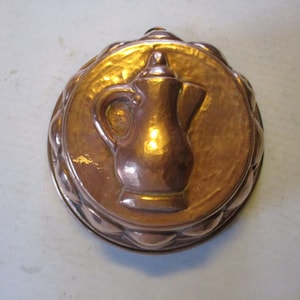 May include: A vintage copper mold with a pitcher design. The round mold has a decorative scalloped edge and a hammered texture. The pitcher is in the center of the mold and has a handle and spout.