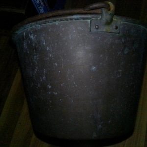 May include: A weathered copper bucket with a dark, oxidized patina and visible surface spots. The bucket features a sturdy metal handle attached to the rim with riveted brackets, showing signs of age and wear across its cylindrical body.
