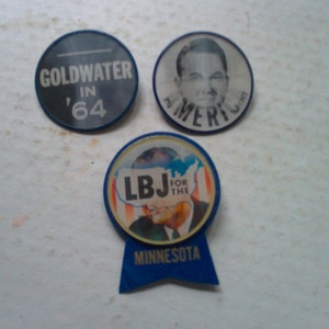 May include: Three vintage political campaign buttons. The first button is blue and white with the text "Goldwater in '64". The second button is blue and white with the text "For America" and a picture of a man. The third button is blue and white with the text "LBJ for the" and a picture of a man. The button has a blue ribbon with the text "Minnesota".