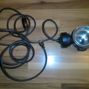 May include: A black electrical cord with a plug on one end and a vintage-style spotlight on the other end. The spotlight has a round, silver reflector and a black metal housing.